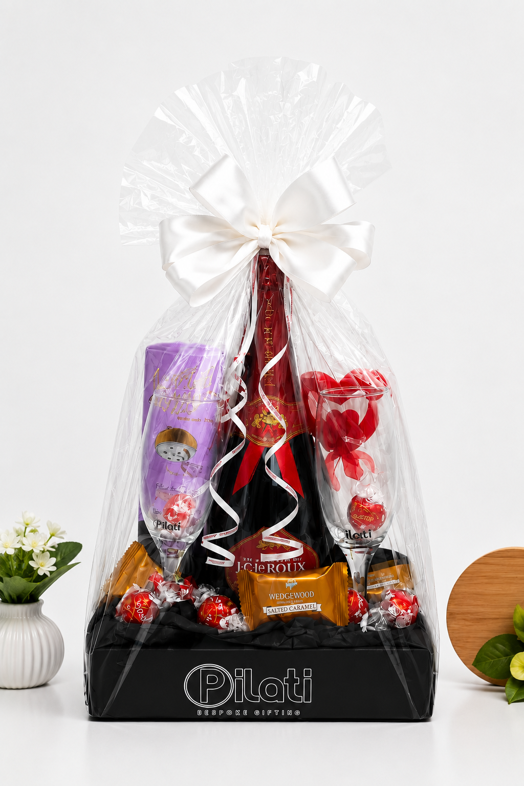 A luxury chocolate and gift set in a black box branded 'Pilati Gourmet Gifting', wrapped in clear cellophane with a large white bow. The set includes a bottle of Valtori sparkling wine, a purple cup of espresso macchiato, heart-shaped chocolates, and assorted chocolates on a white background.