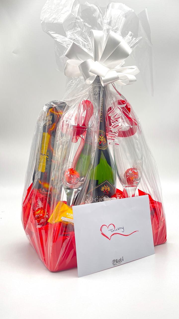 A Valentine's Day gift basket wrapped in clear cellophane with a white bow, containing a bottle of red wine, chocolates, and a small card that reads 'Love xOXo'.
