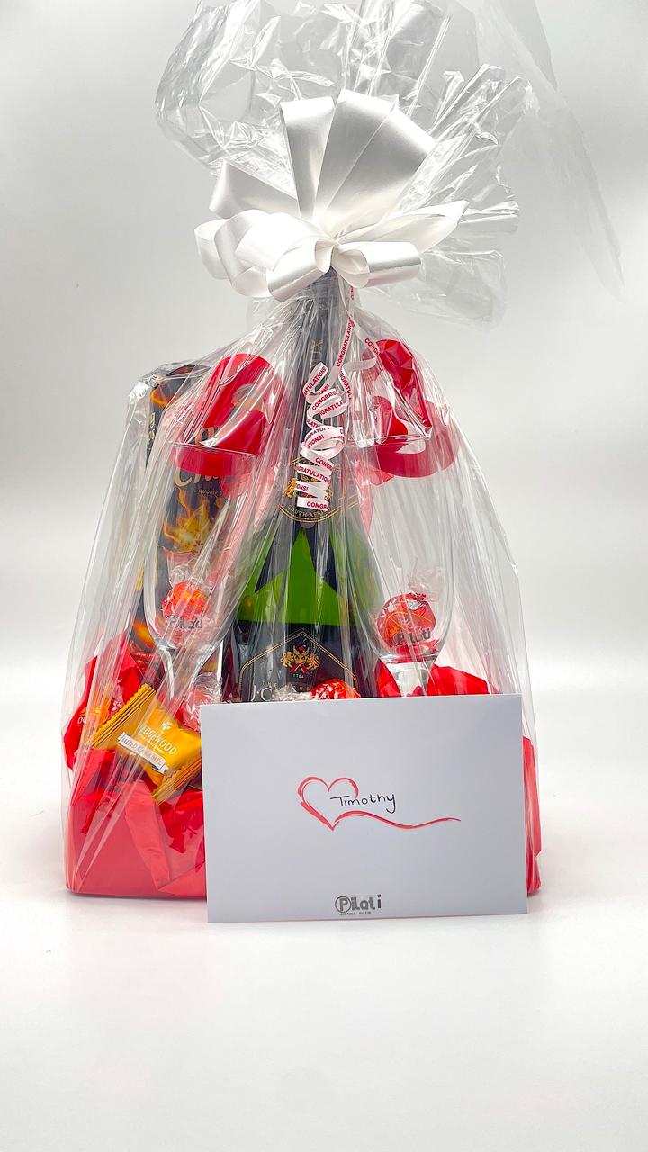 A Valentine's Day gift basket wrapped in clear cellophane with a large white bow, filled with red chocolates, candy canes, and a green bottle, with a white card in front that reads 'Happy Valentine's Day' and has the brand 'Pasi' printed at the bottom.