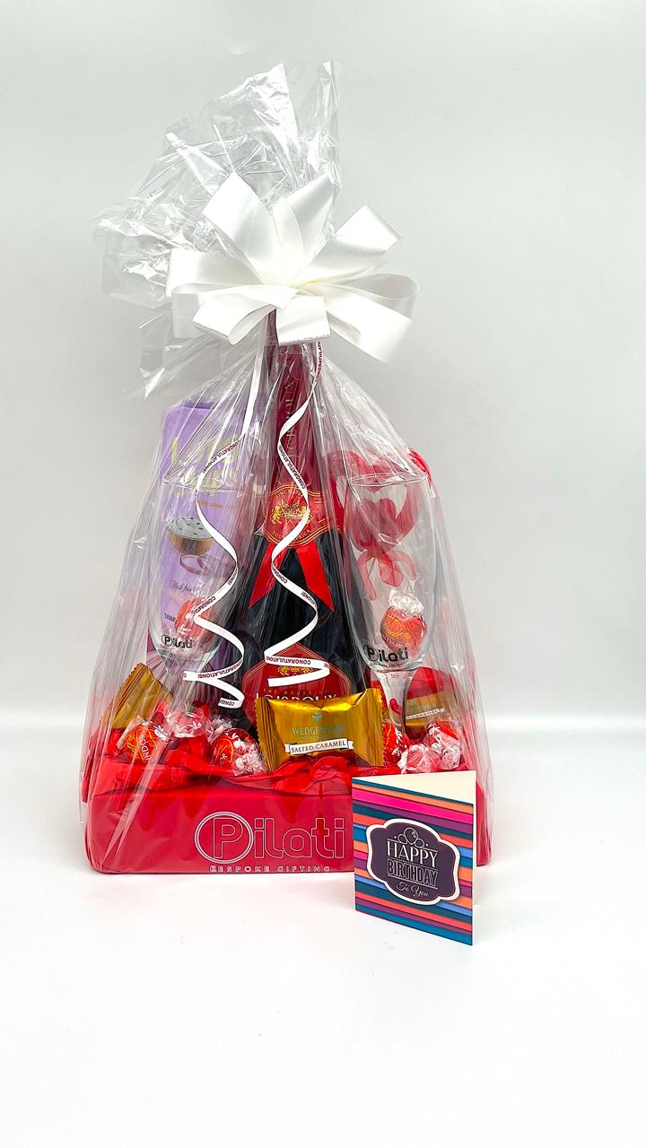 A packaged gift basket filled with assorted candies and chocolates, wrapped in clear cellophane with a large white bow on top. A red base box is visible at the bottom, and a small striped card reading 'Happy' is placed in front.