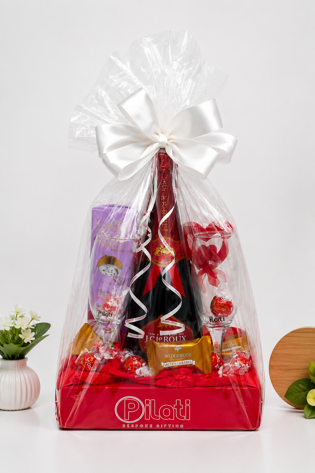 A Valentine's Day gift basket from Pilati featuring a bottle of red wine, chocolates, and treats, wrapped in clear love with a large white bow on a white background.