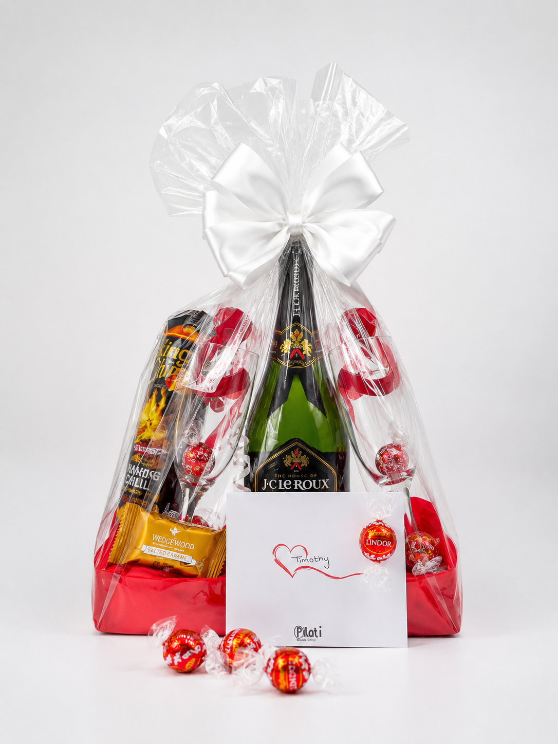 A champagne gift basket wrapped in clear cellophane with a large white bow, containing a bottle of J.C.