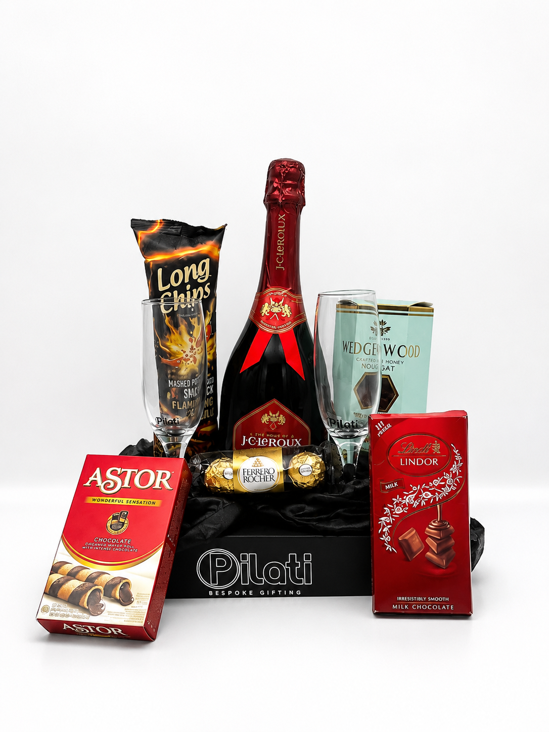 A curated gift basket or hamper on a white studio background, featuring a bottle of Jérévols champagne, two champagne flutes, a box of Long Chips, an Astor chocolate box, a Lindor chocolate box, and Ferrero Rocher chocolates arranged in a black basket with a Pilati logo at the bottom.