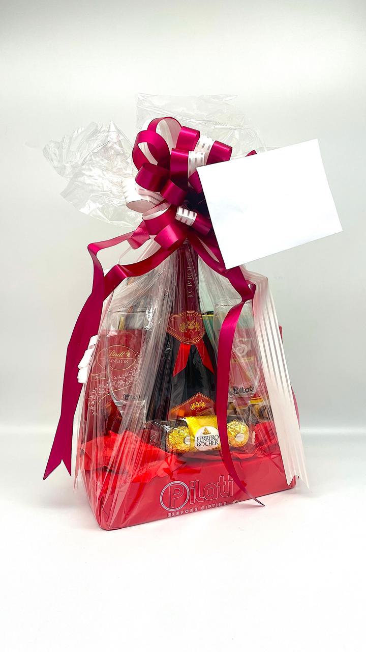 A gift basket packaged in clear love with a large magenta ribbon and bow, containing assorted packaged snacks and treats, with a blank white gift tag attached, set against a plain white background.