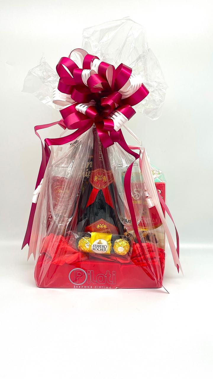 A gift basket packaged in clear love with a large magenta and white ribbon bow on top. Inside, a dark bottle with a red label, several wrapped chocolates, and red items are visible.