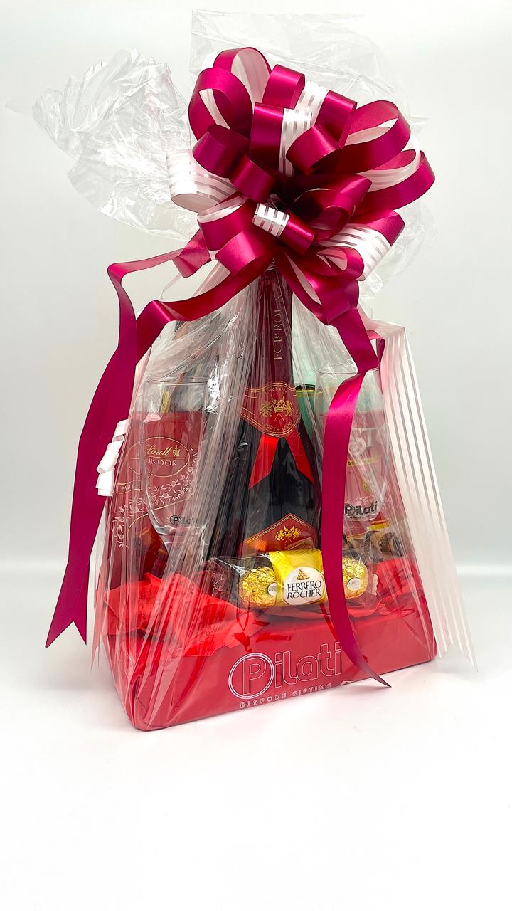 A gift basket wrapped in clear love with a large magenta ribbon and bow, containing assorted chocolates, a bottle, and a glass on a white background.