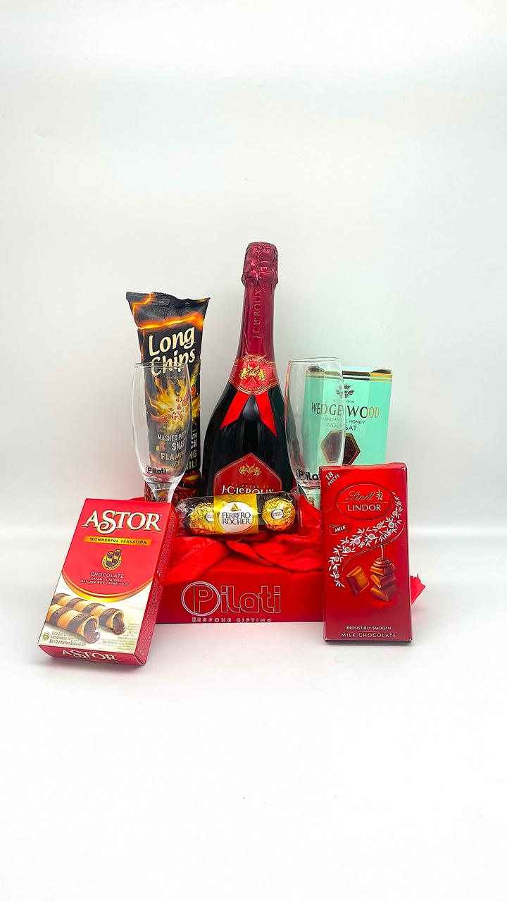 A gift basket arrangement featuring a bottle of champagne, a Long Choc chocolate bar, a can of Red Wing wine, an Astor chocolate box, and a red decorative basket with a red bow, set against a plain white background.