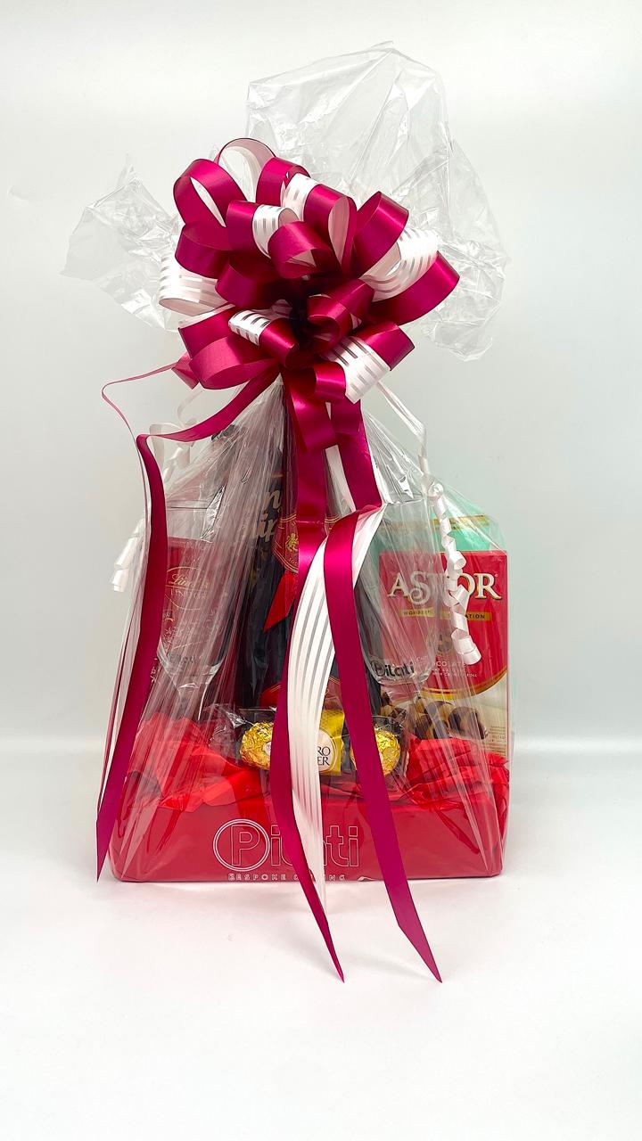 A gift basket wrapped in clear love with a large magenta and white ribbon bow on top, containing assorted chocolates and packaged treats, set against a plain white background.