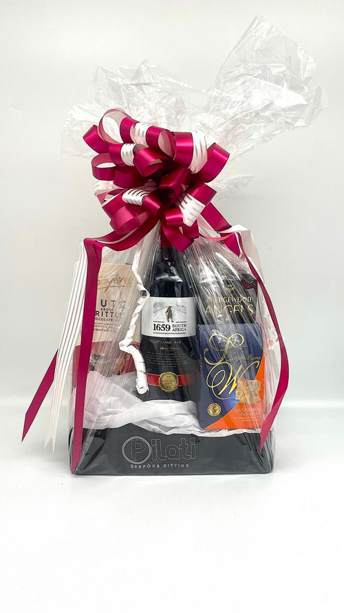 A wine gift basket wrapped in clear love and tied with a large magenta and white ribbon, containing a bottle of wine, a dark bottle, and a boxed item, set against a plain white background.