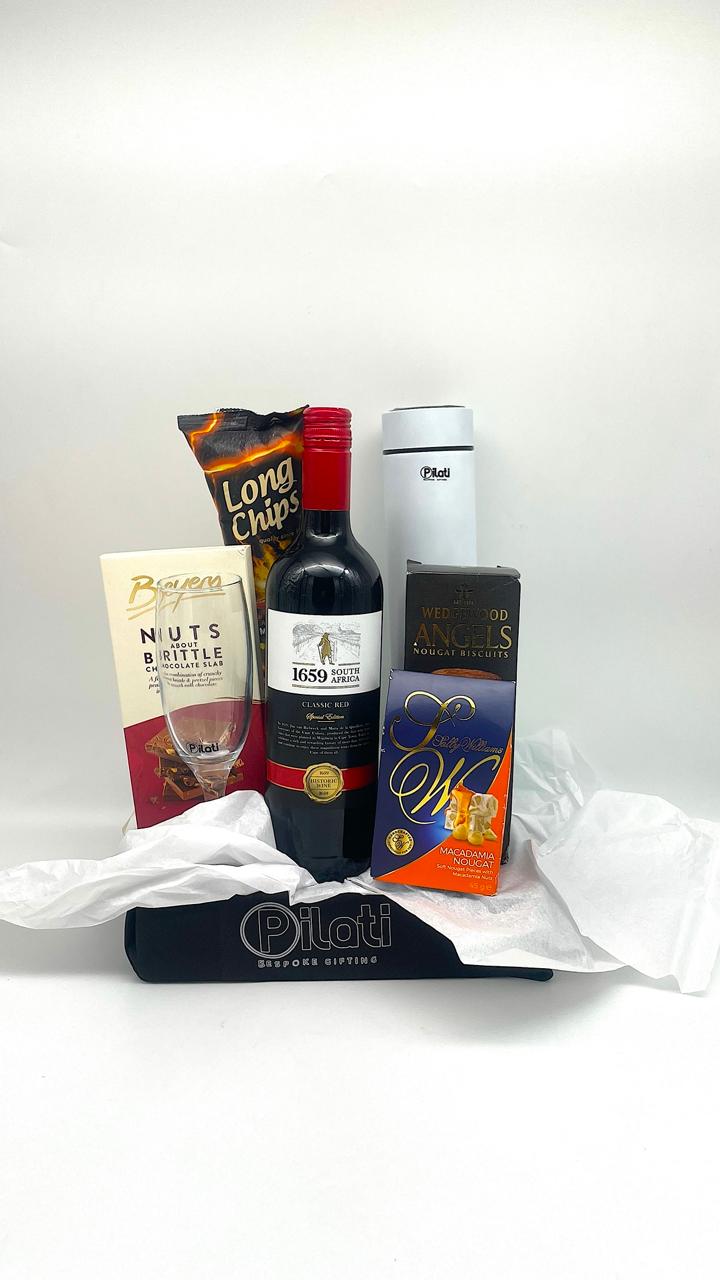 A gift basket or curated food and beverage selection on a white background, including a bottle of red wine, a bag of Long Chips, a box of Big, a white insulated cup, and a dark blue box, arranged with white tissue paper and a black cloth base.