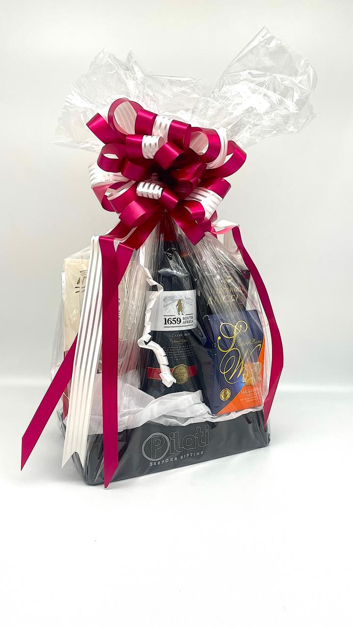 A gift basket wrapped in clear love with a large magenta and white ribbon bow on top, containing assorted items including a dark bottle, a white box, and a small orange item, set against a plain white background.