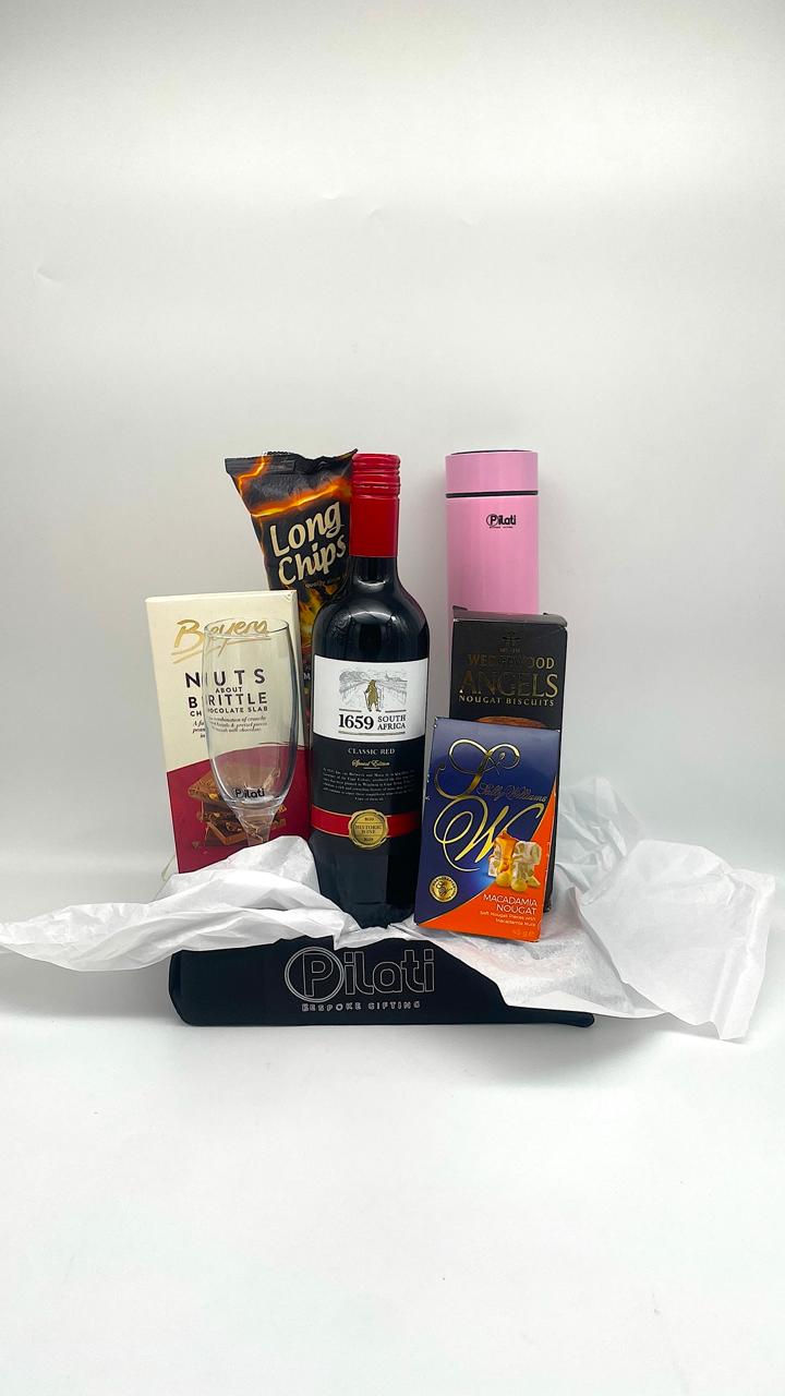 A curated gift basket or hamper containing a bottle of red wine, a bag of Long Chips, , a pink thermos, and a blue and orange decorative box, arranged on white tissue paper inside a black fabric basket with a 'Plot' label.