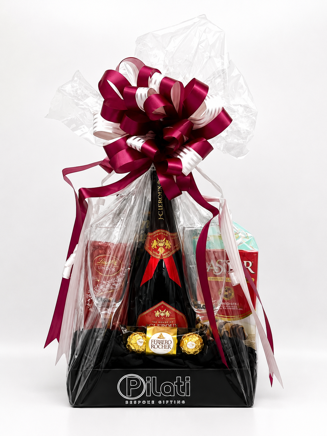 A luxury gift basket from Pilotti Resinace Gifting, featuring a bottle of wine, chocolates, and other gourmet items wrapped in clear LOVE and tied with a large dark red and white ribbon, set against a plain white background.